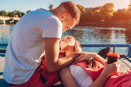 女人接吻男友幸福情侣坐在河边, 拥抱亲吻.躺在男友膝上的女人照片